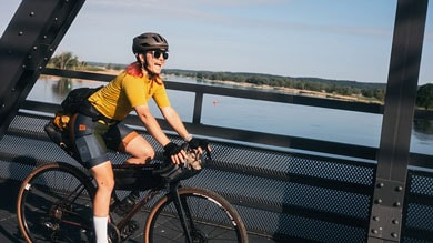 Eine Radfahrerin fährt mit einem Gravelbike über eine Brücke, gekleidet in ein gelbes Trikot, schwarze Radhose und Helm. Das Rad hat mehrere Taschen, was auf eine lange Tour hindeutet.