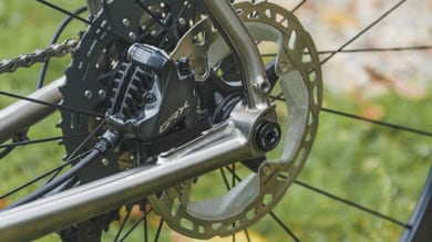 Detaillierte Ansicht der hinteren Scheibenbremse und des Antriebs eines Fahrrads, mit Shimano GRX-Hydraulikbremse und einer Kassette mit mehreren Gängen. Der Titanrahmen bildet einen Kontrast zu den schwarzen Komponenten.