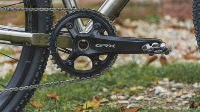 Nahaufnahme eines Shimano GRX-Kurbelsatzes an einem Gravelbike, mit einem einzelnen Kettenblatt und einem SPD-Pedal. Im Hintergrund sind Gras und herabgefallene Herbstblätter zu sehen.