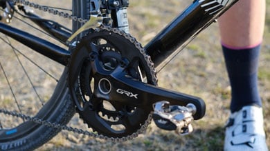 Ein Shimano GRX-Kurbelset an einem schwarzen Gravelbike, ein Fahrer mit weißen Radschuhen steht daneben.