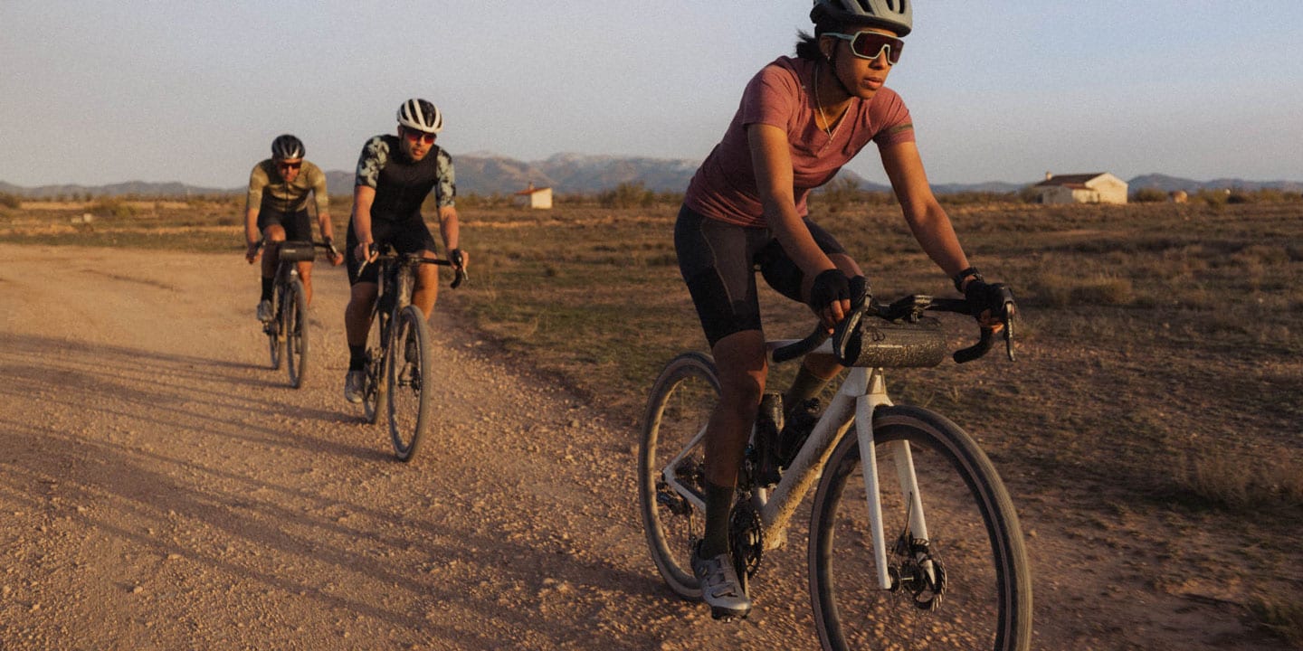 Drei Radfahrer fahren mit Gravelbikes auf einem staubigen Weg während der goldenen Stunde. Im Hintergrund sind Berge und kleine Häuser zu sehen. Der vordere Fahrer trägt ein pinkes Trikot und schwarze Shorts.