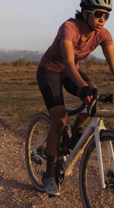 Drei Radfahrer fahren mit Gravelbikes auf einem staubigen Weg während der goldenen Stunde. Im Hintergrund sind Berge und kleine Häuser zu sehen. Der vordere Fahrer trägt ein pinkes Trikot und schwarze Shorts.
