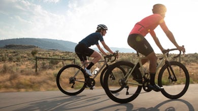 Two cyclists riding on a road in a rural landscape with mountains in the background