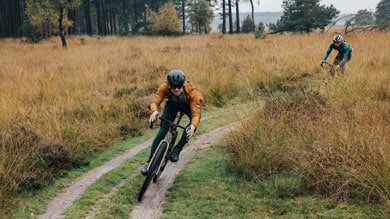 Dwóch rowerzystów jadących polną ścieżką przez zalesiony obszar z wysoką trawą.