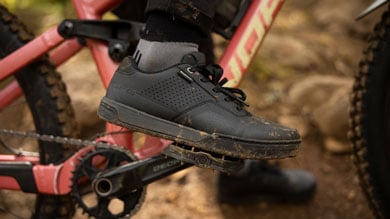 Person's foot on a mountain bike pedal wearing a muddy black cycling shoe