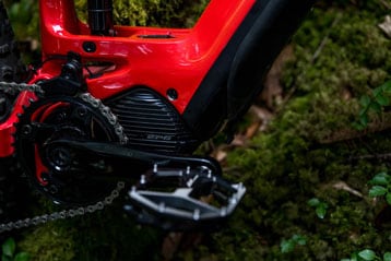 Close-up of an electric bike's red frame, motor, chain, and pedal over mossy ground.