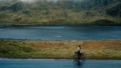 Cyclist rides along a calm lake with fog-covered mountains in the background