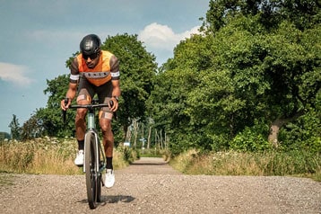 Fietser in oranje trui rijdt op een grindpad omringd door bomen.