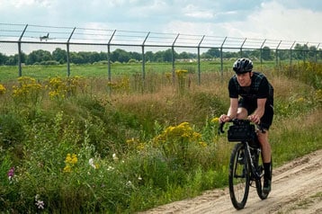 Fietser rijdt op een onverhard pad nabij een omheind veld met een vliegtuig in de lucht.
