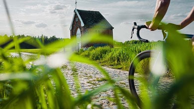 Fietser rijdt over een kasseipad richting een kleine bakstenen kapel omgeven door groen.