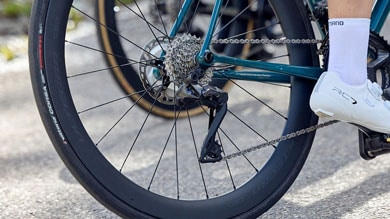 Close-up of a cyclist's rear bike wheel, chain, and derailleur on a road bike.