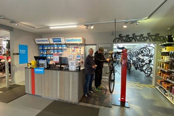 Personen stehen an einem Serviceschalter in einem Fahrradladen mit ausgestellten Fahrradteilen.