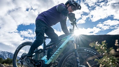 Mountainbikerin in lila Jacke fährt bergauf auf einem Canyon-Fahrrad, während die Sonne durch den Rahmen scheint und Berge im Hintergrund zu sehen sind