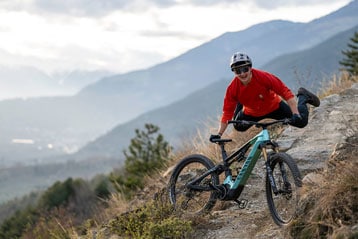 Lächelnder Mountainbiker im roten Trikot macht ein Wheelie auf einem Canyon-Fahrrad auf einem felsigen Pfad mit Tal und Bergen in der Ferne