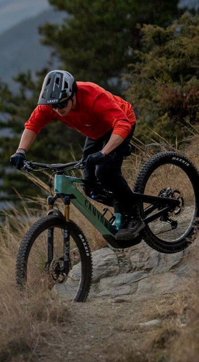 Mountainbiker im orangefarbenen Trikot fährt ein Canyon-Fahrrad auf einem felsigen Bergpfad mit dramatischen Wolken und Gipfeln im Hintergrund