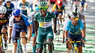 Ciclistas no sprint para a linha de chegada de uma prova de estrada, com um deles celebrando energicamente.