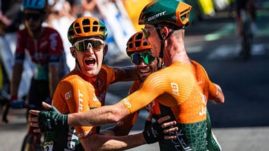 Drie wielrenners in oranje truien vieren met een vrolijke omhelzing tijdens een race.