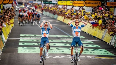 Dos ciclistas celebran mientras cruzan la línea de meta en una carrera, con fans animando de fondo