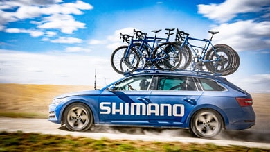 Blue Shimano car with bicycles on roof rack driving on a rural road under a partly cloudy sky