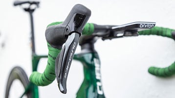 Close-up of a bicycle's handlebar and gear shifter with green handlebar tape and text 'Dura-Ace' and 'Cube'