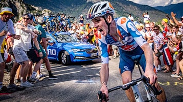 Cyclist in blue and white gear racing uphill with cheering crowd and support vehicle in the background