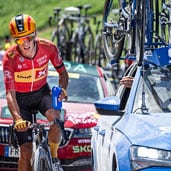 Ciclista con maillot rojo sosteniendo un bidón azul al lado de un vehículo de apoyo durante una carrera