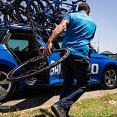 Person in Shimano attire carrying a bicycle wheel next to a blue car loaded with bikes