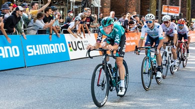Eine Gruppe von Radrennfahrern auf einer Stadtstraße, angeführt von einem Fahrer im grünen Trikot. Zuschauer jubeln hinter Absperrungen mit Bannern, darunter Shimano- und UCI-Logos.