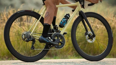 Nahaufnahme der Beine eines Radfahrers auf einem cremefarbenen Rennrad mit schwarzen Rädern. Eine Trinkflasche ist am Rahmen befestigt, der Fahrer trägt schwarze Socken und Radschuhe.