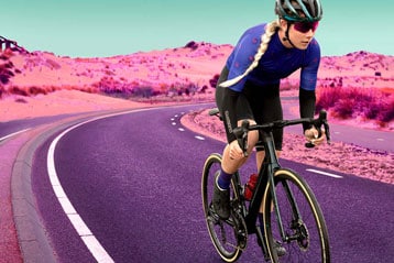Eine Radfahrerin in blauem Trikot und schwarzer Hose fährt ein schwarzes Rennrad auf einer kurvigen Asphaltstraße. Der Hintergrund zeigt digital veränderte rosa Sanddünen unter einem türkisfarbenen Himmel.