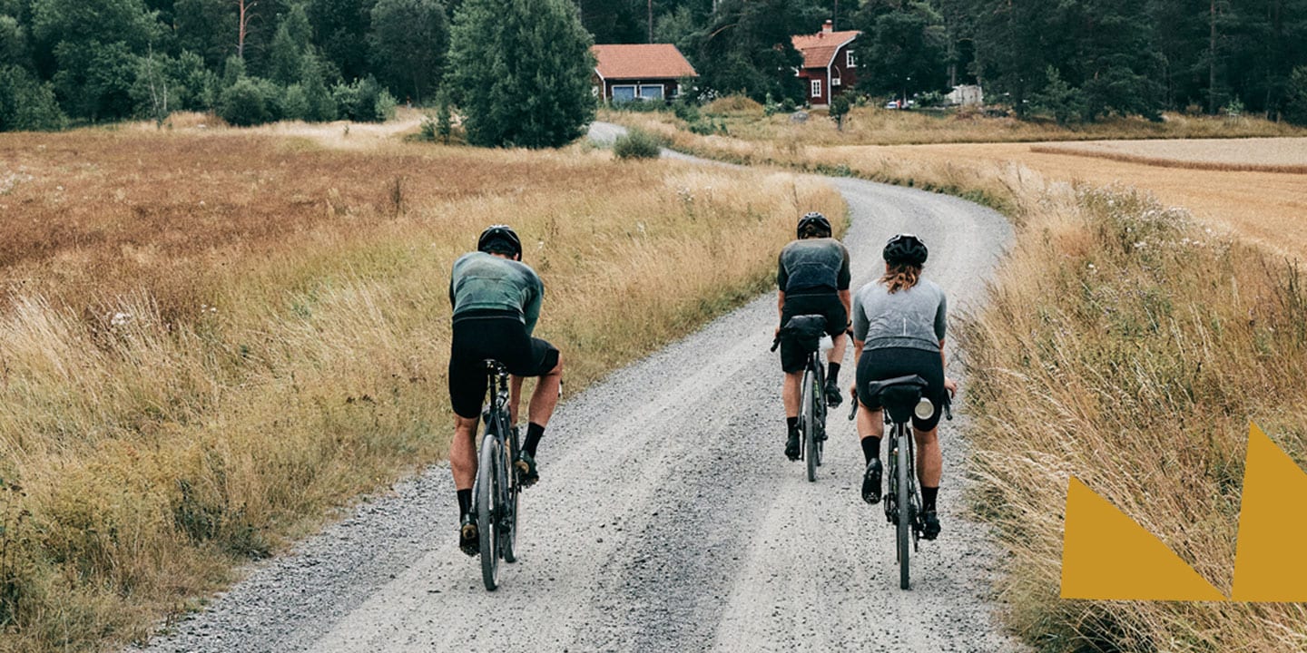 Three cyclists riding on a gravel road through a rural landscape with fields and trees.