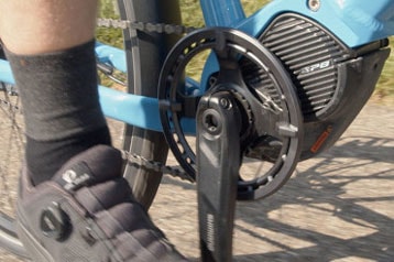 Nahaufnahme eines Fahrradschuhs auf einem Pedal, mit sichtbarer Kurbel und Motor.
