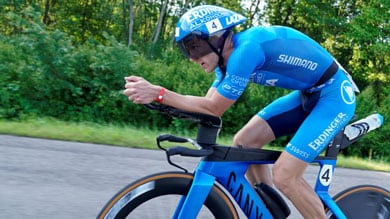 Eine Nahaufnahme eines Radfahrers im blauen Skinsuit und Helm auf einem Zeitfahrrad. Er fährt auf einer Straße mit grünen Bäumen und hält eine aerodynamische Position.