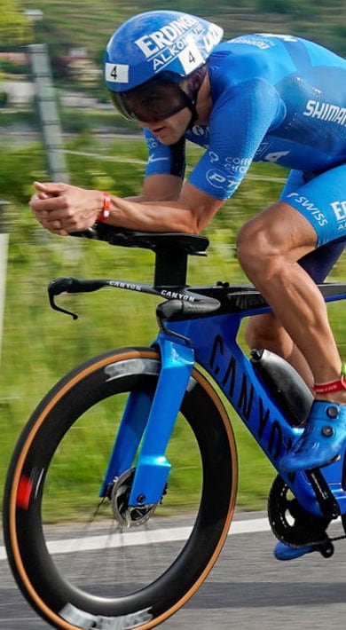 Ein Profiradfahrer im blauen Skinsuit fährt ein blaues Zeitfahrrad auf einer Landstraße. Der verschwommene Hintergrund mit Grünflächen und Häusern deutet auf hohe Geschwindigkeit hin.
