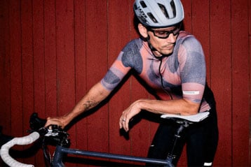 Cyclist in a helmet and sunglasses leaning on a bicycle against a red wooden wall