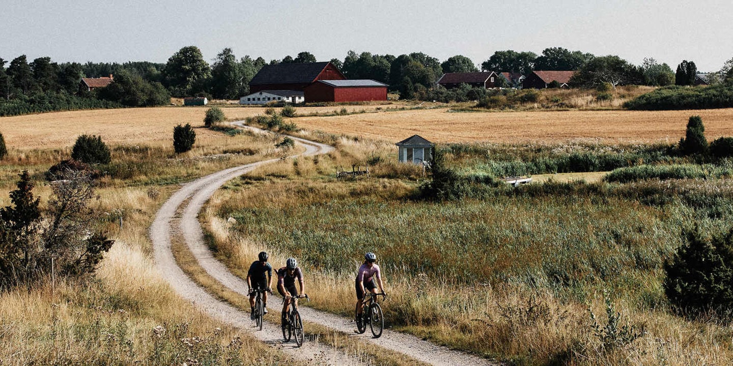 Three cyclists riding on a dirt path through a rural landscape with fields and farm buildings