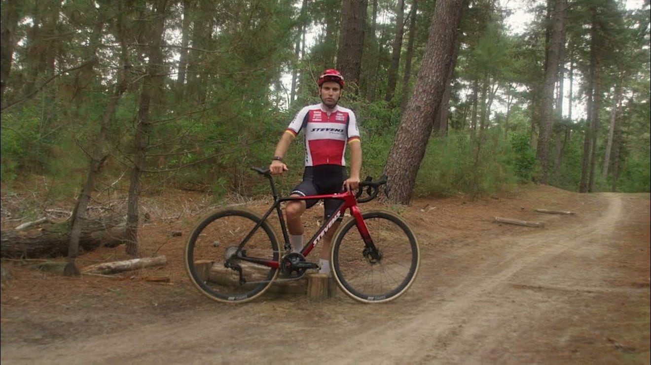 Ein Profiradsportler im roten und weißen Stevens-Trikot steht in einem Wald und posiert mit seinem rot-schwarzen Fahrrad auf einem sandigen Weg, umgeben von hohen Bäumen.