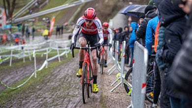 Ein entschlossener Radfahrer kämpft sich durch ein schlammiges Cyclocross-Rennen, fährt auf einem nassen Kiesweg, während Zuschauer hinter Absperrungen stehen.
