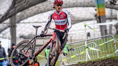 Ein Cyclocross-Fahrer in einem roten und weißen Trikot trägt sein schlammiges Fahrrad eine steile Böschung hinauf, während Zuschauer hinter Absperrungen zuschauen.