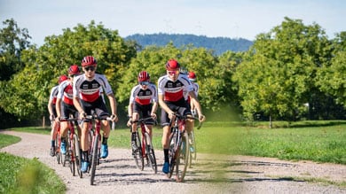 Eine Gruppe von Radfahrern in roten und weißen Trikots fährt in Formation auf einem Schotterweg, umgeben von üppigem Grün und Bäumen unter klarem blauem Himmel.