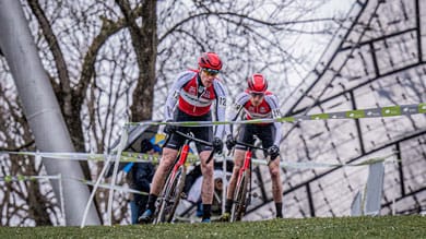 Zwei Radfahrer mit roten Helmen und weißen Trikots rasen durch eine schlammige Cyclocross-Strecke, ihre Gesichter voller Konzentration, während sie eine schwierige Passage bewältigen.