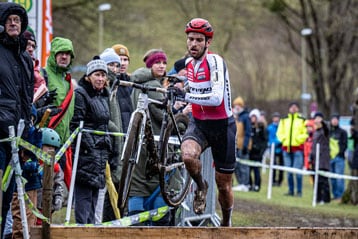 Ein entschlossener Cyclocross-Athlet im schlammigen roten und weißen Trikot springt über eine Holzhürde, während er sein schmutzbedecktes Fahrrad trägt, während Zuschauer in warmer Kleidung das Rennen verfolgen.