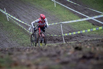 Ein Radfahrer mit rotem Helm und weißem Trikot kämpft sich einen schlammigen Cyclocross-Hang hinauf, hält den Lenker fest und navigiert durch das unebene Gelände.
