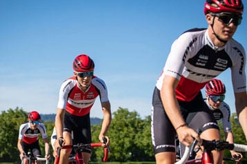 Eine Gruppe von Radfahrern in passenden rot-weiß-schwarzen Trikots fährt an einem sonnigen Tag auf einem Kiesweg, umgeben von grünen Bäumen und blauem Himmel.