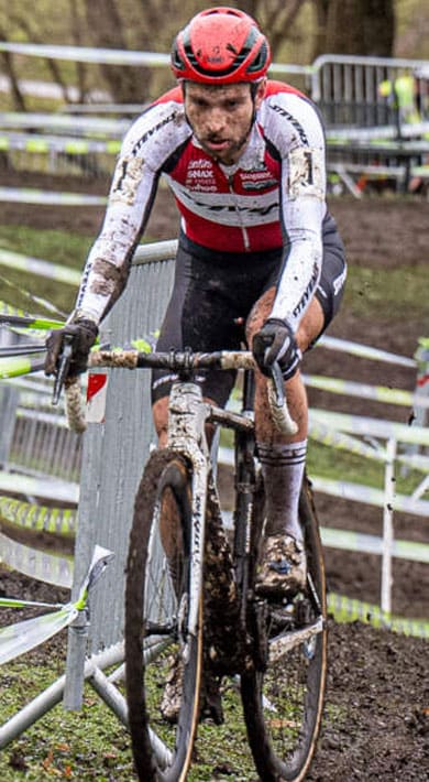 Ein schlammiger Cyclocross-Fahrer im weißen und roten Trikot fährt durch eine kurvenreiche, mit Absperrbändern gesäumte Strecke und kämpft sich durch das nasse Gelände, während Zuschauer hinter den Absperrungen zuschauen.