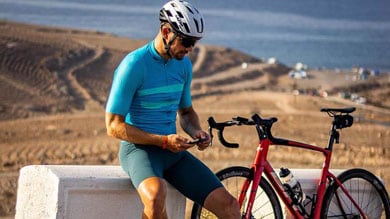 Fietser in blauwe outfit zit bij een rode fiets en kijkt op zijn telefoon, met een mooi uitzicht op de achtergrond.