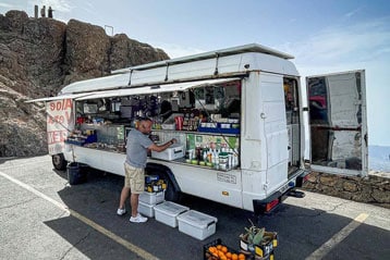 Man staat bij een witte bestelwagen foodtruck met voorraden, geparkeerd op een bergweg onder een heldere blauwe lucht.
